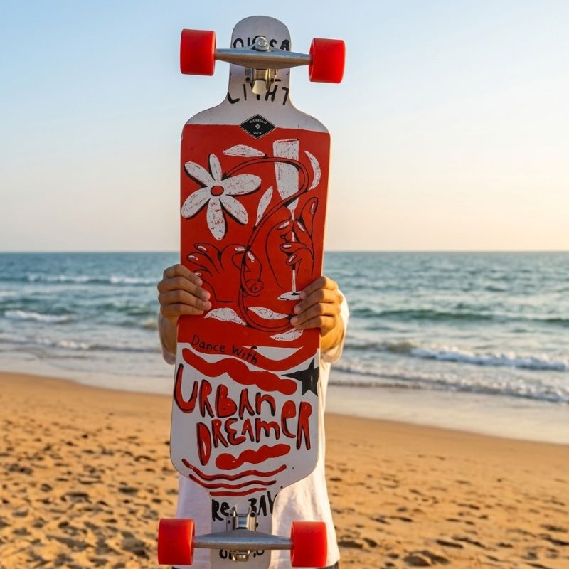 A longboard skateboard with a red and white graphic design featuring a flower, face illustration, and the text 'URBAN DREAMER' and 'Dance No. 4', held by a person standing on a sandy beach with the ocean in the background.